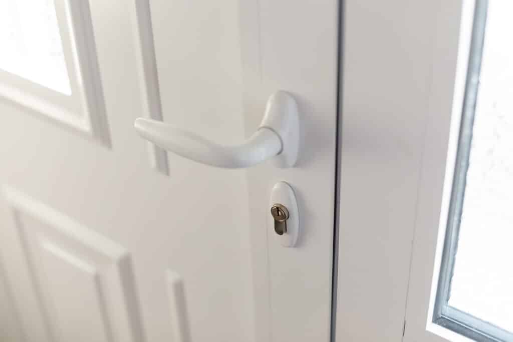 Poignée de porte blanche et serrure : gros plan intérieur Gros plan sur une poignée de porte blanche et une serrure cylindrique sur une porte intérieure moulurée, près d'une fenêtre lumineuse.