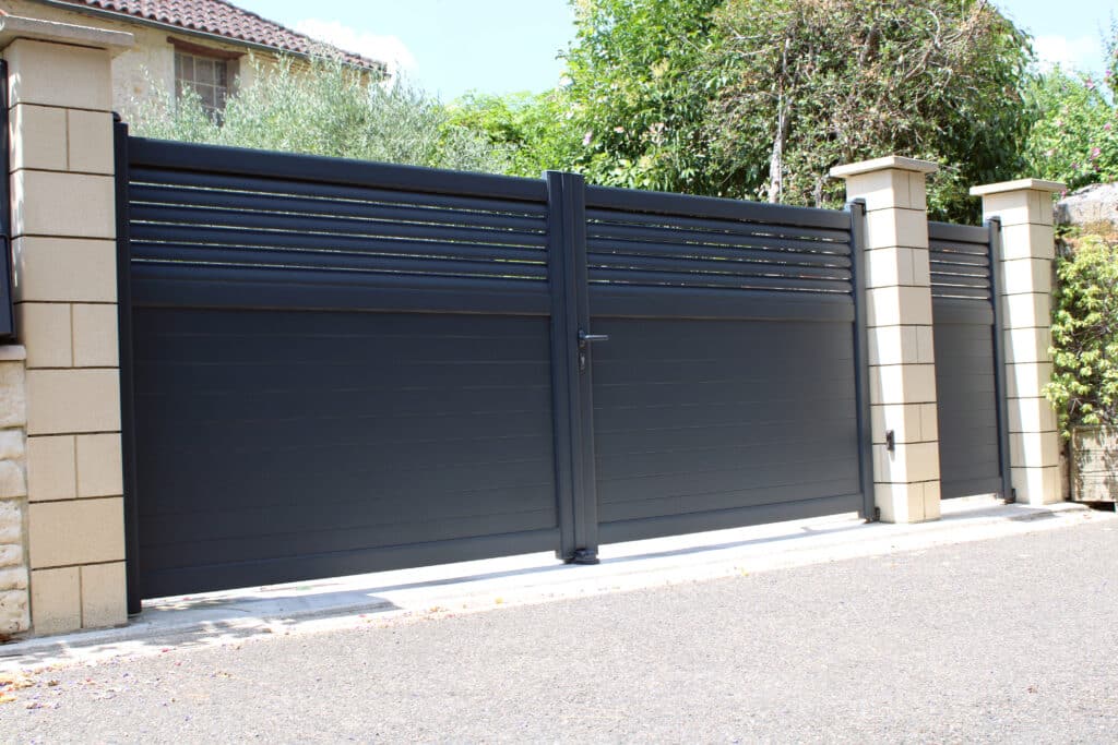 Portail Coulissant Moderne Anthracite et Piliers Pierre Portail coulissant moderne anthracite avec lames horizontales, soutenu par des piliers en pierre claire, devant une maison avec végétation luxuriante.