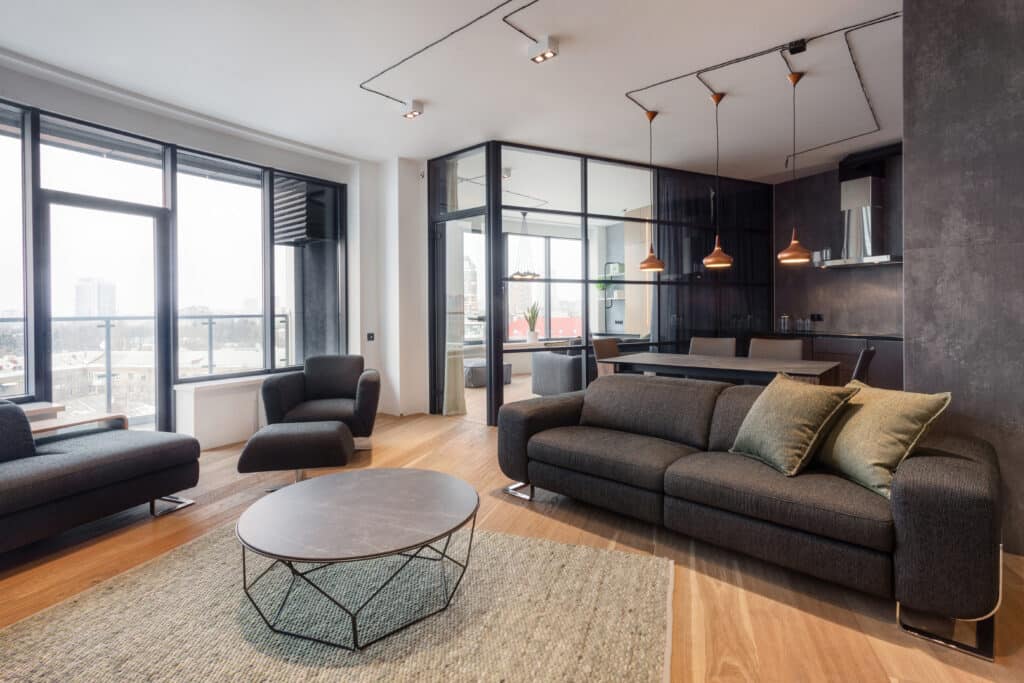 Salon Moderne Design : Canapé, Verrière et Cuisine Intégrée Salon moderne avec canapé gris foncé, fauteuil, table basse ovale à piètement géométrique, et vue sur une cuisine/salle à manger à travers une cloison vitrée noire.